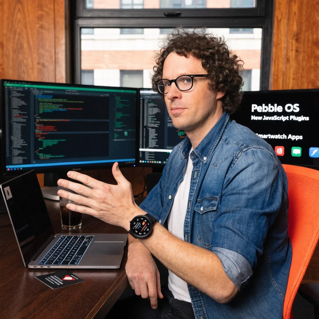 A realistic photo of Eric Migicovsky demonstrating Pebble OS with new JavaScript plugins and smartwatch apps on a Pebble smartwatch.