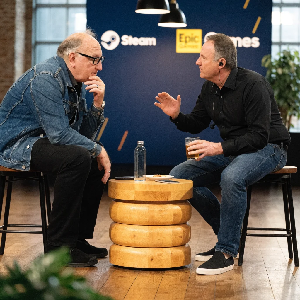 A candid photo of Gabe Newell and Tim Sweeney discussing AI-driven game development at a tech conference with Steam and Epic Games logos in the background.