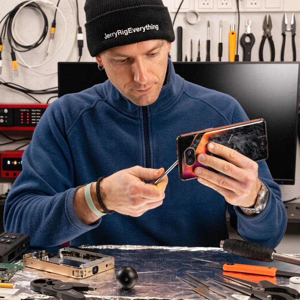 A realistic photo showing the Trump Mobile T1 smartphone undergoing scratch and bend tests by JerryRigEverything expert Zack Nelson, with HTC U24 Pro components and iFixit teardown tools in the background.