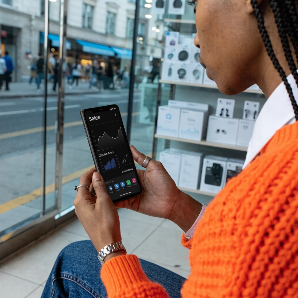 A realistic photo of a Samsung Galaxy Z Fold 8 smartphone held by a person in a tech store, with a display showing sales graphs and market share data.