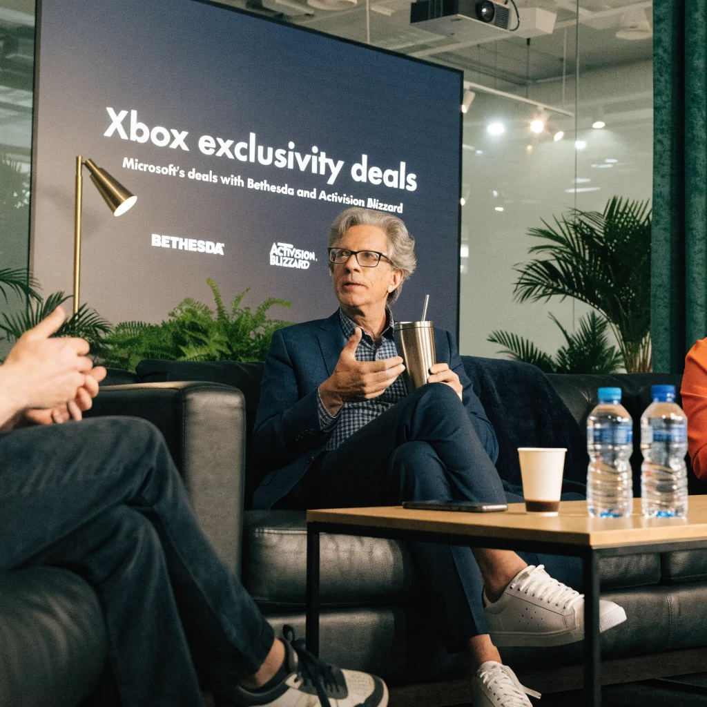 A realistic photo of Shawn Layden discussing Microsoft's Xbox exclusivity deals with Bethesda and Activision Blizzard at a gaming industry conference.